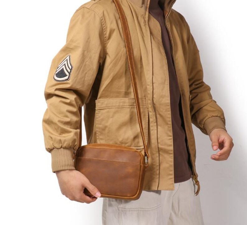 Puede incluir: Un bolso bandolera de cuero marr&oacute;n con forma rectangular y una correa larga y ajustable. La persona lleva una chaqueta color beige con un parche de estilo militar en la manga. El bolso tiene un dise&ntilde;o sencillo y cl&aacute;sico.