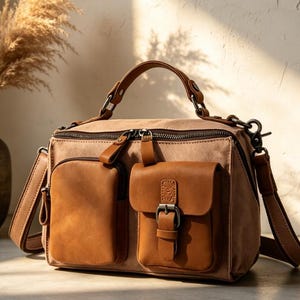 May include: A tan canvas and brown leather handbag with two front pockets and a top handle. The bag has a zippered closure and a detachable shoulder strap. The leather pockets have a buckle closure. The bag is sitting on a light-coloured surface.