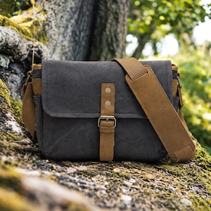 May include: A charcoal grey messenger bag featuring tan leather-like accents, a front flap with a vertical strap, a metal buckle, and two silver-toned rivets. The bag includes a wide, adjustable tan shoulder strap with a padded section for comfort.