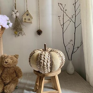 Op de afbeelding: Een decoratieve pompoen gemaakt van geweven natuurlijke vezels en beige stof, tentoongesteld op een kleine houten kruk. De pompoen heeft een steel en is omgeven door een neutrale achtergrond. Een teddybeer is zichtbaar op de achtergrond.