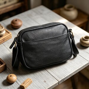 Puede incluir: Un bolso bandolera de cuero negro con forma rectangular y cremallera superior. El bolso tiene un bolsillo frontal y una correa larga ajustable. Los herrajes son dorados. El bolso est&aacute; sobre una superficie de madera.