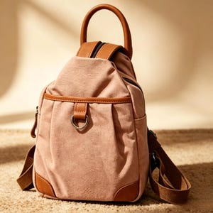 Könnte beinhalten: Ein rosafarbenes Canvas-Rucksack mit braunen Lederakzenten. Die Tasche hat einen oberen Griff, verstellbare Schultergurte und eine Vordertasche mit einem Metallring. Die Ecken und der Besatz sind mit braunem Leder verstärkt.