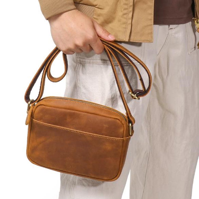 Puede incluir: Un bolso bandolera de cuero marr&oacute;n con forma rectangular y un bolsillo frontal. El bolso tiene cierre de cremallera y una correa larga y ajustable. El bolso es sostenido por una persona.