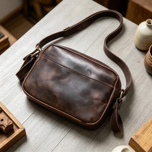 Puede incluir: Un bolso bandolera de cuero marr&oacute;n con una correa larga ajustable. El bolso tiene forma rectangular con un bolsillo frontal y herrajes dorados. El cuero tiene un aspecto envejecido y texturizado.