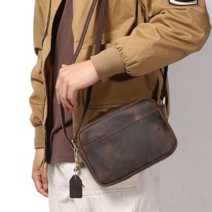 Puede incluir: Un bolso bandolera de cuero marr&oacute;n con forma rectangular y una correa larga. El bolso tiene un acabado envejecido y una peque&ntilde;a etiqueta adjunta a la cremallera. El bolso se lleva al hombro.