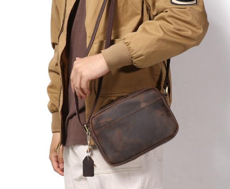 Puede incluir: Un bolso bandolera de cuero marr&oacute;n con forma rectangular y una correa larga. El bolso tiene un acabado envejecido y una peque&ntilde;a etiqueta adjunta a la cremallera. El bolso se lleva al hombro.