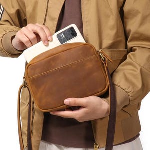 Puede incluir: Un bolso bandolera de cuero marr&oacute;n con forma rectangular y una correa larga y ajustable. El bolso tiene cierre de cremallera y un bolsillo frontal. Una persona est&aacute; metiendo un smartphone blanco en el bolso.