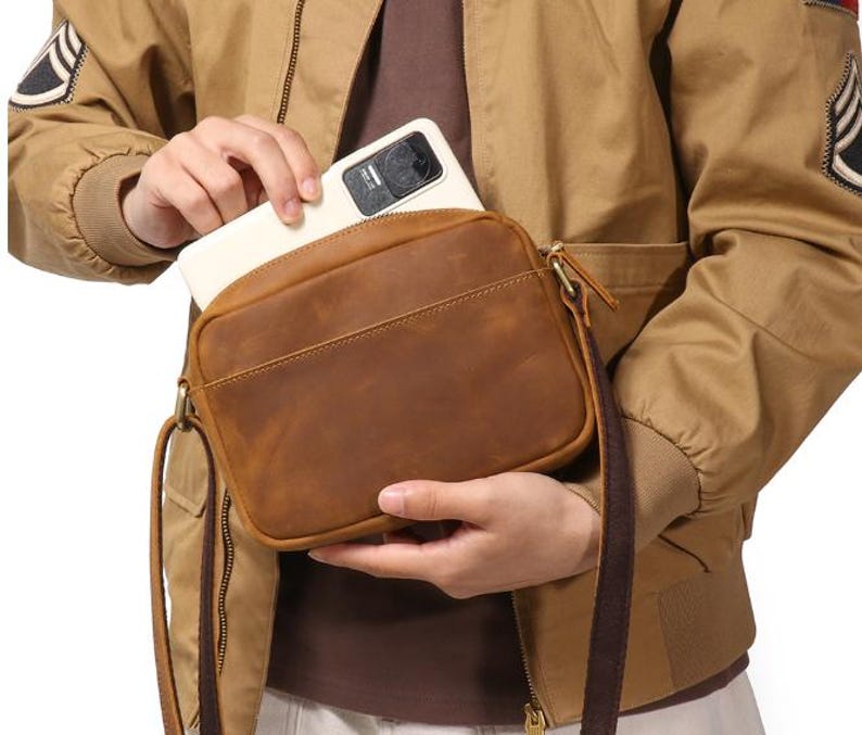 Puede incluir: Un bolso bandolera de cuero marr&oacute;n con forma rectangular y una correa larga y ajustable. El bolso tiene cierre de cremallera y un bolsillo frontal. Una persona est&aacute; metiendo un smartphone blanco en el bolso.