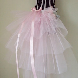 Puede incluir: Una falda de tul rosa con un diseño de capas y largas cintas de satén rosa. La falda está en un maniquí y tiene un corsé a rayas blancas y negras debajo.