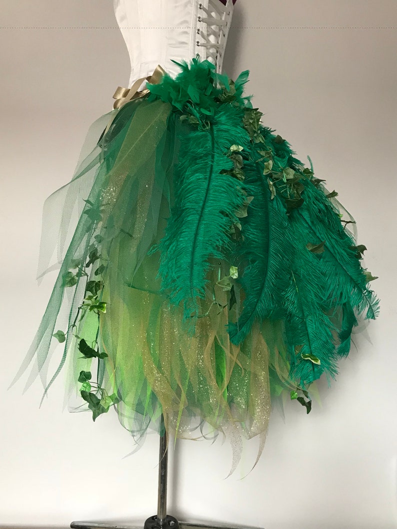 May include: A green tulle skirt with green feathers and green leaves. The skirt is layered with gold glitter tulle.