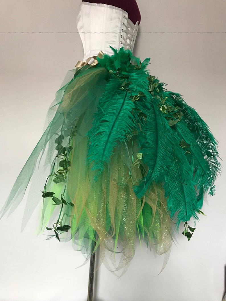 May include: A green and gold tulle skirt with green feathers and ivy accents. The skirt is layered and has a sparkly finish.