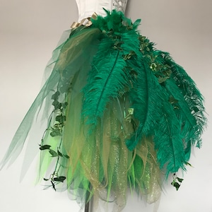 May include: A green and gold tulle skirt with green feathers and ivy accents. The skirt is layered and has a sparkly finish.