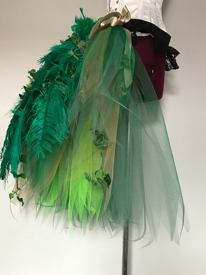 May include: A green, gold, and lime green tulle skirt with green feathers and ivy accents. The skirt is layered and has a flowing, whimsical design.