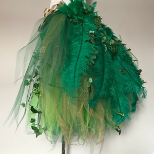 May include: A green tulle skirt with green feathers and green leaves. The skirt is layered with gold glitter tulle.