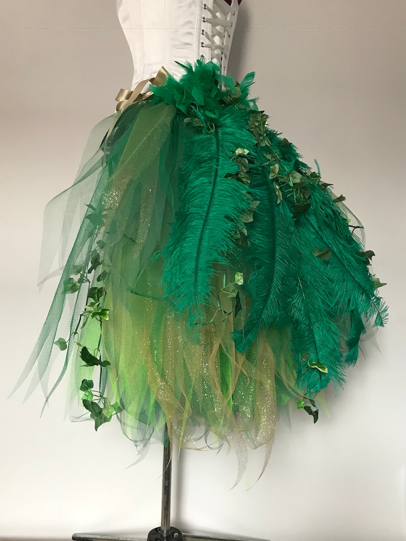 May include: A green tulle skirt with green feathers and green leaves. The skirt is layered with gold glitter tulle.