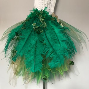 May include: A green tulle skirt with green feathers and green leaves. The skirt is on a white dress form.