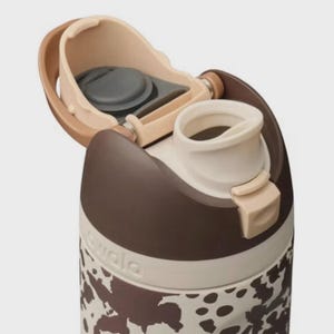 May include: Close-up of a brown and white water bottle with a cow print design. The bottle has a flip-top lid with a beige and brown color scheme. The brand name "owala" is visible on the side of the bottle.