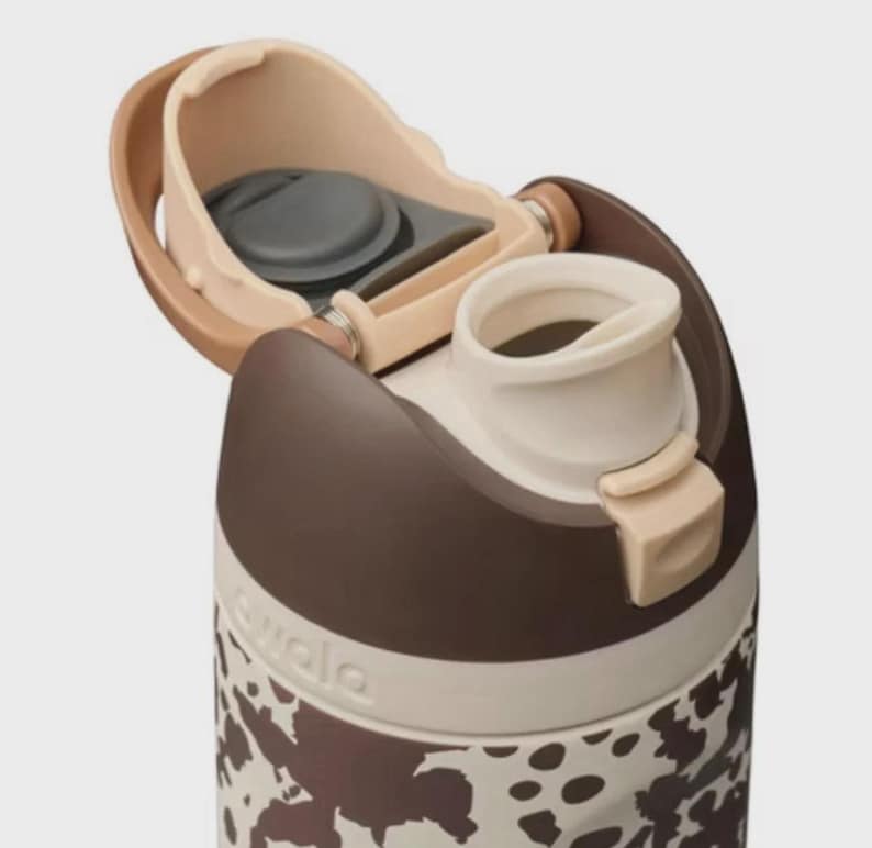 May include: Close-up of a brown and white water bottle with a cow print design. The bottle has a flip-top lid with a beige and brown color scheme. The brand name "owala" is visible on the side of the bottle.