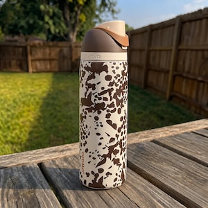 May include: A white and brown patterned water bottle with a brown lid and handle. The bottle has the word "owala" printed on it. The bottle is sitting on a wooden surface.