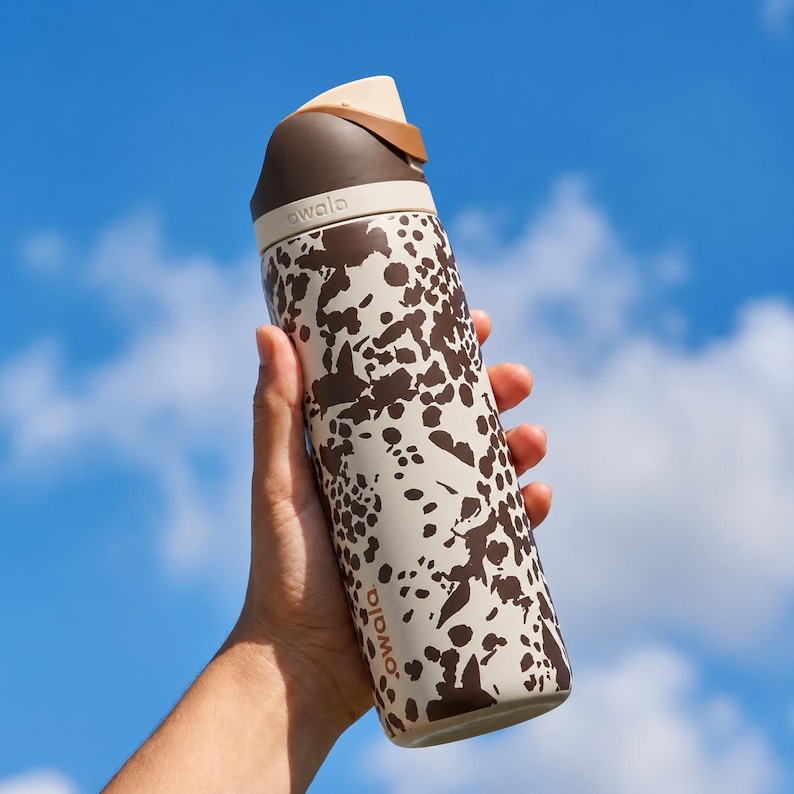 May include: A stainless steel water bottle with a brown and cream spotted pattern is held against a blue sky. The bottle has a brown lid and a cream-colored top. The brand name "owala" is printed on the bottle.