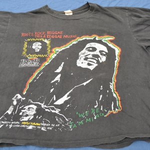 K&ouml;nnte beinhalten: Schwarzes T-Shirt mit einem wei&szlig;en Bild von Bob Marley. Das Shirt tr&auml;gt den Text "Roots Rock, Reggae This a Reggae Music" und "We're Jamming".