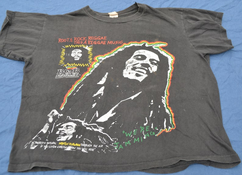 K&ouml;nnte beinhalten: Schwarzes T-Shirt mit einem wei&szlig;en Bild von Bob Marley. Das Shirt tr&auml;gt den Text "Roots Rock, Reggae This a Reggae Music" und "We're Jamming".