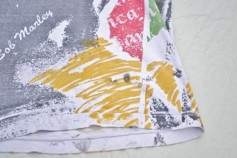 May include: A white t-shirt with a colorful abstract print featuring yellow, red, green, and gray paint splatters. The text "Bob Marley" is printed in black on the shirt.
