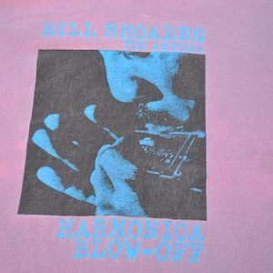 May include: A vintage purple t-shirt with a black and white image of a person playing a harmonica. The text "Bill Rhoades 7th Annual Harmonica Blow-Off" is printed on the shirt.