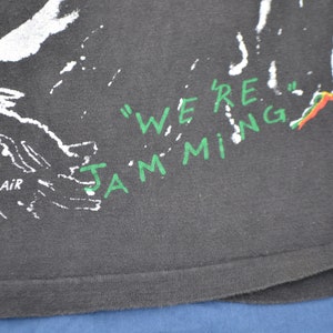 K&ouml;nnte beinhalten: Schwarzes T-Shirt mit wei&szlig;em und gr&uuml;nem Text, der "We're Jamming" lautet, und einem gr&uuml;n-gelb-roten Design. Der Text "Through the air you will hear" ist ebenfalls sichtbar.