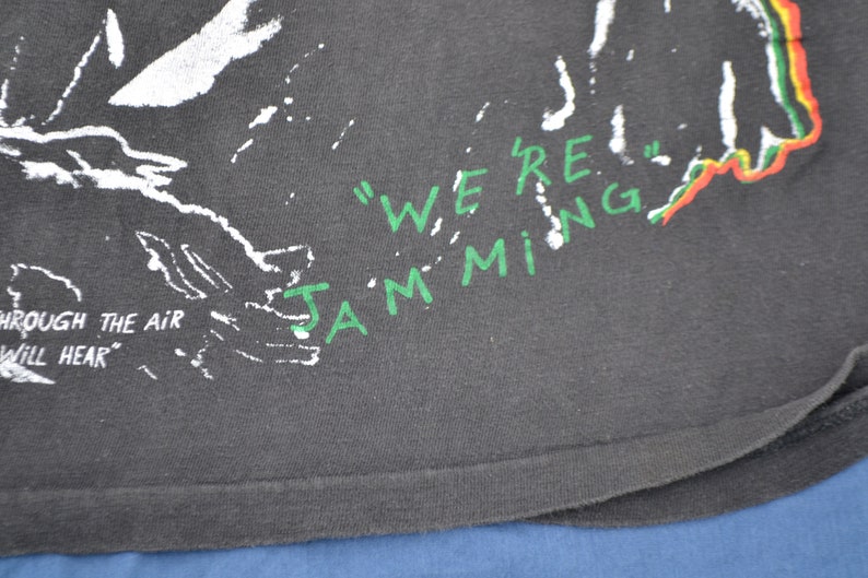 K&ouml;nnte beinhalten: Schwarzes T-Shirt mit wei&szlig;em und gr&uuml;nem Text, der "We're Jamming" lautet, und einem gr&uuml;n-gelb-roten Design. Der Text "Through the air you will hear" ist ebenfalls sichtbar.