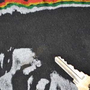 K&ouml;nnte beinhalten: Schwarzes T-Shirt mit einem wei&szlig;en Grafikdruck eines Gesichts und einem roten, gelben und gr&uuml;nen Streifen oben.
