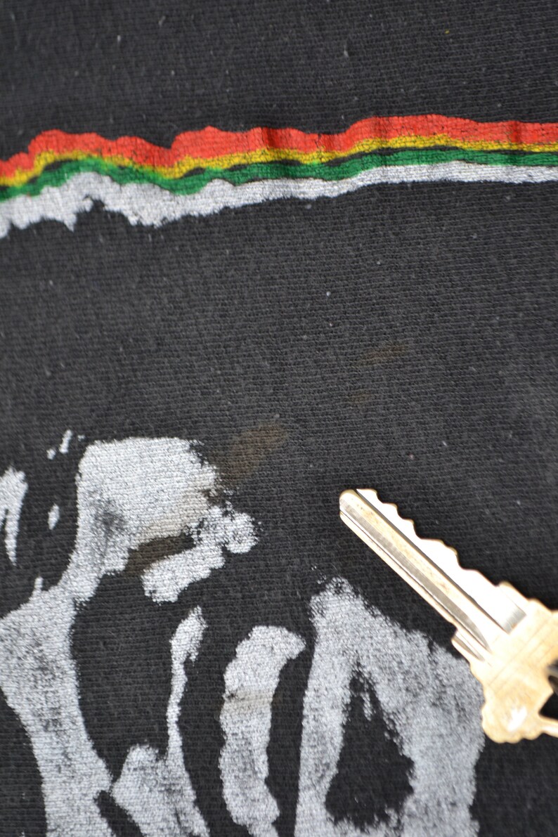 K&ouml;nnte beinhalten: Schwarzes T-Shirt mit einem wei&szlig;en Grafikdruck eines Gesichts und einem roten, gelben und gr&uuml;nen Streifen oben.
