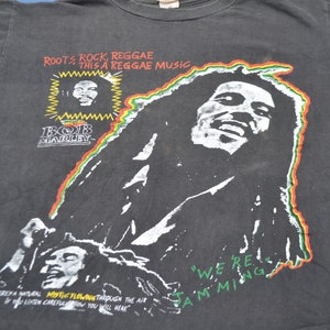K&ouml;nnte beinhalten: Schwarzes T-Shirt mit einem Grafikdruck von Bob Marley. Der Druck zeigt ein Portr&auml;t von Bob Marley mit dem Text "Roots Rock, Reggae This a Reggae Music" und "We're Jamming".