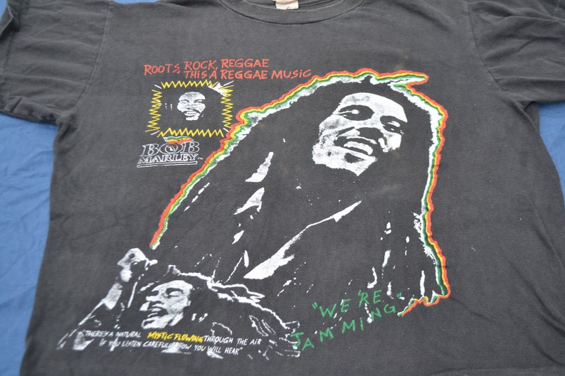 K&ouml;nnte beinhalten: Schwarzes T-Shirt mit einem Grafikdruck von Bob Marley. Der Druck zeigt ein Portr&auml;t von Bob Marley mit dem Text "Roots Rock, Reggae This a Reggae Music" und "We're Jamming".