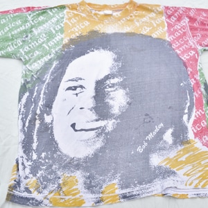 May include: A white t-shirt with a colorful print of Bob Marley's face and the words "Bob Marley" on the front. The shirt has a green, yellow, and red color scheme.