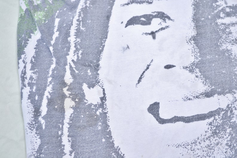 May include: Close-up of a white t-shirt with a black and white graphic print of a person's face. The print is faded and distressed, giving it a vintage look.