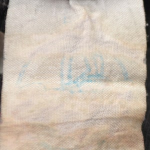 K&ouml;nnte beinhalten: Nahaufnahme eines wei&szlig;en Stoff-Etiketts mit blauer Schrift. Die Schrift ist schwer lesbar.
