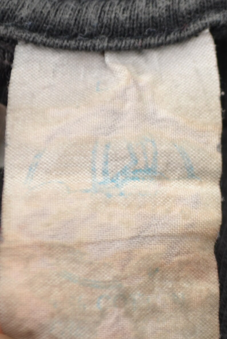 K&ouml;nnte beinhalten: Nahaufnahme eines wei&szlig;en Stoff-Etiketts mit blauer Schrift. Die Schrift ist schwer lesbar.