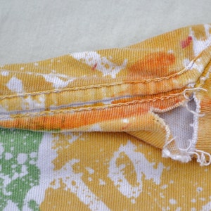 May include: Close-up of a yellow, orange, and white tie-dye fabric with a seam running along the top edge. The fabric has a distressed look with a small tear near the seam.