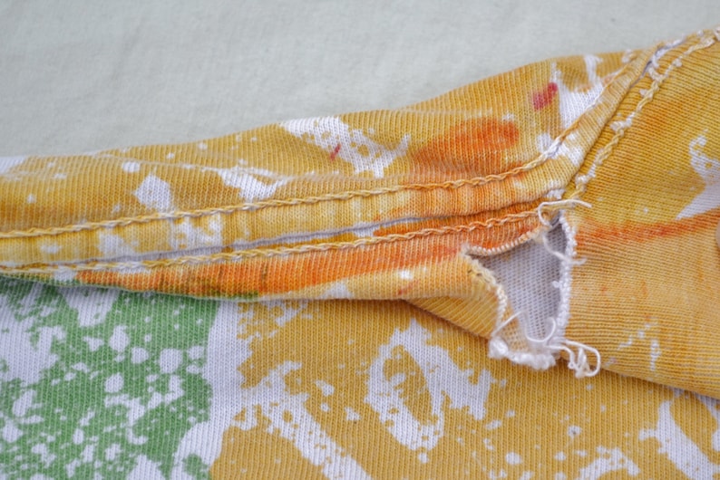 May include: Close-up of a yellow, orange, and white tie-dye fabric with a seam running along the top edge. The fabric has a distressed look with a small tear near the seam.
