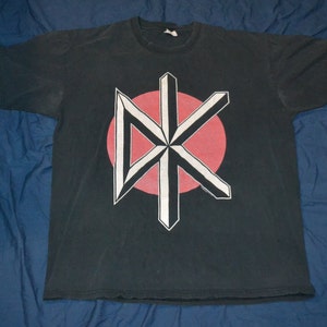 May include: Black t-shirt with a white and red graphic of a stylized symbol. The symbol is a large, geometric star with a red circle in the center.