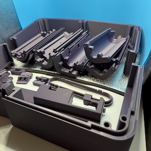 May include: A purple plastic container with several compartments and components. The container is open, revealing various parts, including curved and rectangular pieces. The background is a light blue.