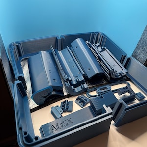 May include: A blue plastic case containing various 3D-printed components. The components include rectangular pieces, brackets, and other geometric shapes. The text "AD5X" is visible on one of the pieces.