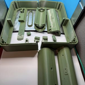 May include: A collection of sage green 3D printed plastic components for an enclosure, including a rectangular frame, curved panels, hinges, and a small rectangular piece with the text AD5X embossed on the surface, all arranged for assembly.