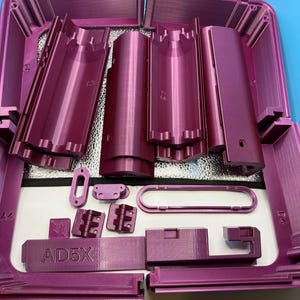 May include: A disassembled, magenta-colored product kit with various components, including cylindrical and rectangular parts, arranged inside a matching case. The text "AD5X" is visible on one of the components. The parts are made of a textured material.