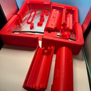 May include: A collection of bright red 3D printed plastic components, including several L-shaped frame pieces, two cylindrical housing segments with screw holes, and various small rectangular and oval connectors arranged on a flat surface.