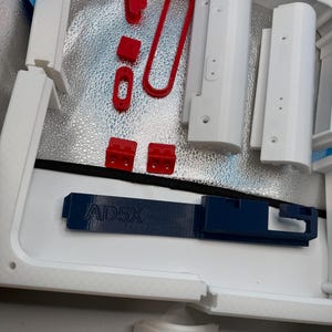 May include: A collection of 3D printed components including white structural frames, red small connectors, and a dark blue horizontal bar with the embossed text AD5X. The parts are arranged on a reflective silver surface within a white enclosure.