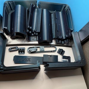 May include: A collection of dark gray 3D-printed components, including rectangular and curved pieces, arranged on a white surface. The text "AD5X" is visible on one of the parts. The components appear to be parts of a larger assembly.