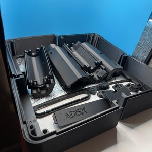 May include: A collection of dark gray 3D printed components arranged inside a rectangular container. The parts include cylindrical housing segments, a flat rectangular piece with the embossed text AD5X, and various small brackets and connectors.