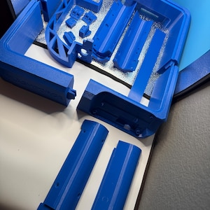 May include: A collection of bright blue 3D-printed components, including rectangular and curved pieces, arranged on a white surface. The parts appear to be from a larger assembly, possibly a housing or enclosure.